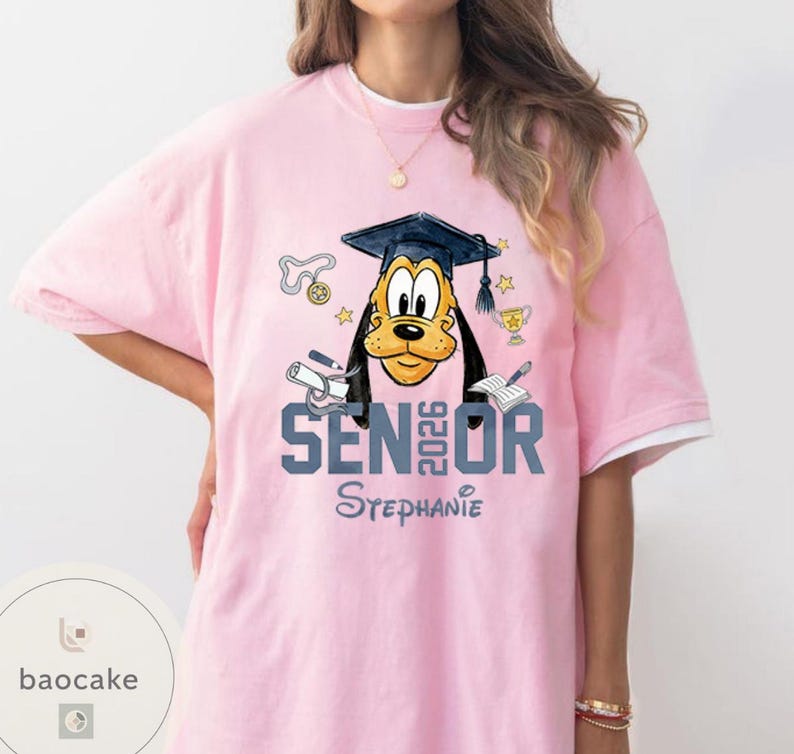 May include: Pink t-shirt with a graduation-themed design featuring a cartoon dog wearing a graduation cap. The shirt says "SENIOR 2026" and "Stephanie". The design includes stars, a trophy, and other graduation-related elements.