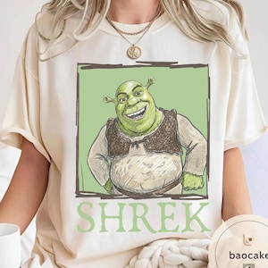May include: Cream-colored t-shirt featuring a smiling green ogre character from the animated film Shrek, set against a light green background within a dark brown frame. The word "SHREK" is printed in green below the image.