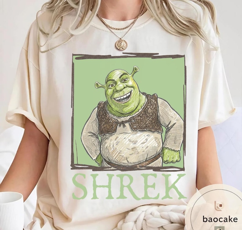 May include: Cream-colored t-shirt featuring a smiling green ogre character from the animated film Shrek, set against a light green background within a dark brown frame. The word "SHREK" is printed in green below the image.