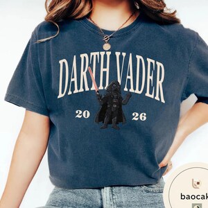 May include: Navy blue t-shirt featuring the text "Darth Vader" in a curved, off-white font. Below the text is a graphic of Darth Vader holding a red lightsaber, with the numbers "20" and "26" on either side. The shirt is a casual style.