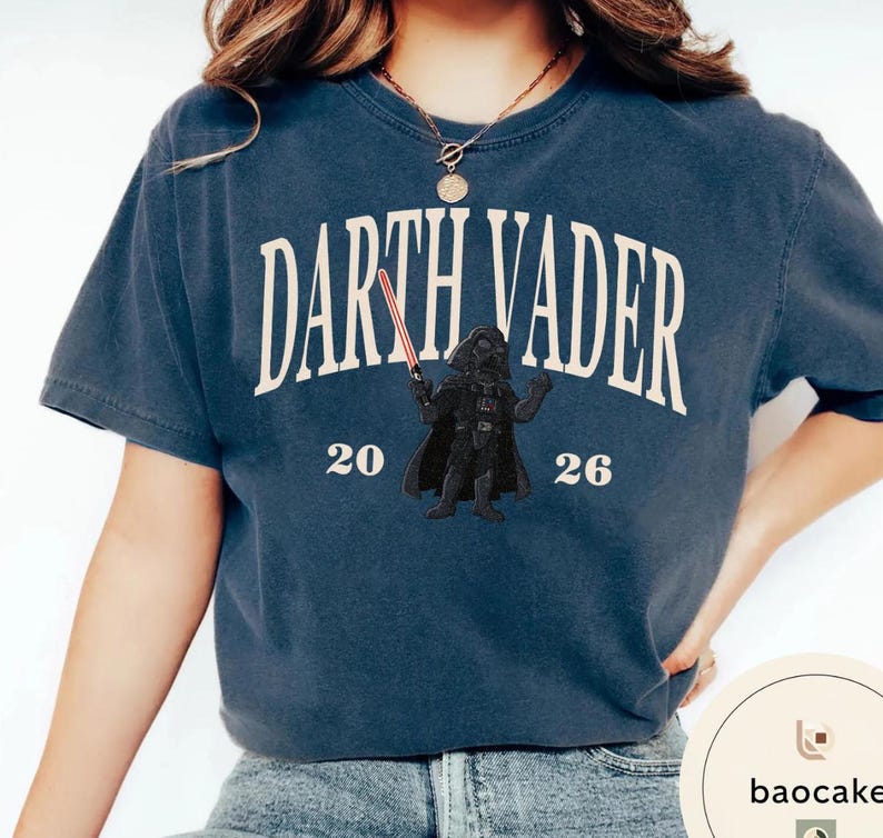 May include: Navy blue t-shirt featuring the text "Darth Vader" in a curved, off-white font. Below the text is a graphic of Darth Vader holding a red lightsaber, with the numbers "20" and "26" on either side. The shirt is a casual style.