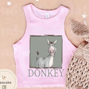 May include: Pink tank top with a cartoon donkey graphic inside a gray rectangle. The word "DONKEY" is printed below the graphic in gray. A tan crocheted item is in the background.