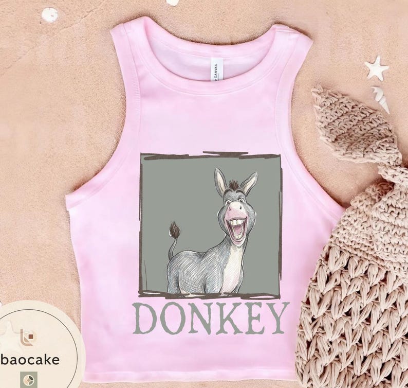 May include: Pink tank top with a cartoon donkey graphic inside a gray rectangle. The word "DONKEY" is printed below the graphic in gray. A tan crocheted item is in the background.