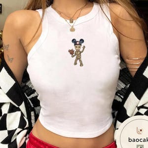 May include: A white sleeveless tank top featuring a small graphic print of a tree-like character wearing blue mouse ears and holding a brown ice cream treat. The top has a high, rounded neckline and a fitted silhouette, crafted from a solid white fabric.