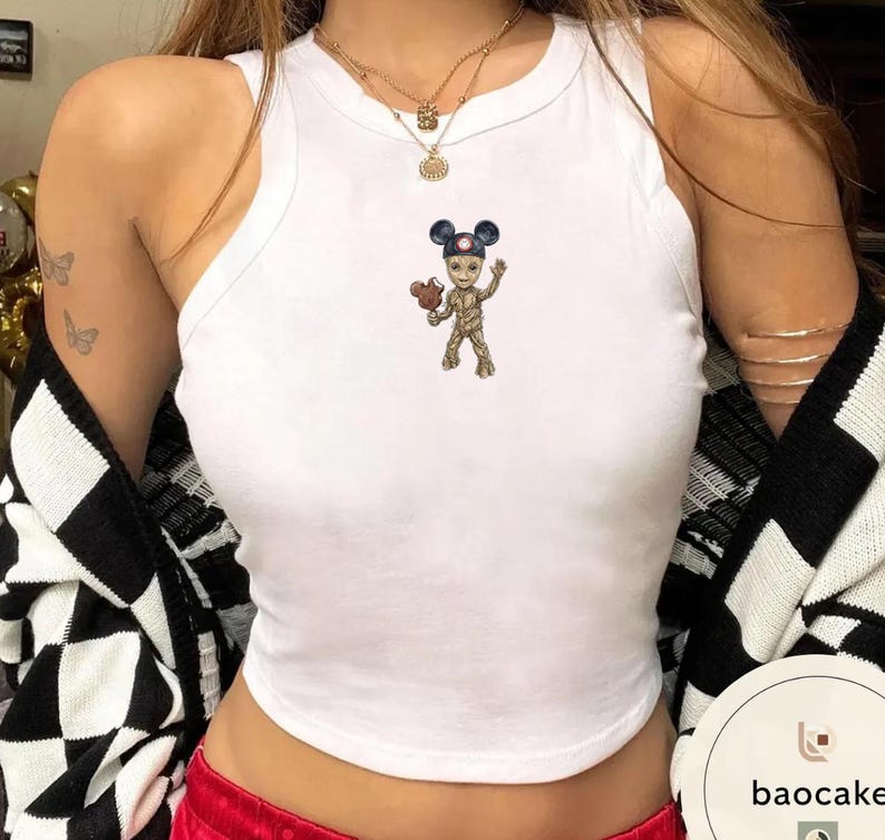 May include: A white sleeveless tank top featuring a small graphic print of a tree-like character wearing blue mouse ears and holding a brown ice cream treat. The top has a high, rounded neckline and a fitted silhouette, crafted from a solid white fabric.