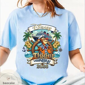 May include: Light blue t-shirt featuring a Disney design with Tigger at the helm of a ship. The design includes the text "Disney", "Tigger Wonder 2026", palm trees, and floral accents. The shirt is a short-sleeved crew neck.
