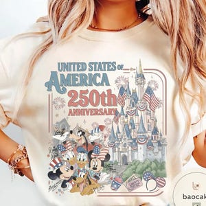 May include: Cream-colored t-shirt featuring a graphic celebrating the 250th anniversary of the United States of America. The design includes a castle, fireworks, and Disney characters like Mickey and Minnie Mouse, all in patriotic colors.