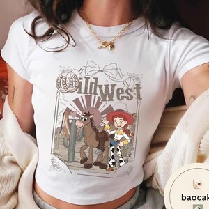 Pu&ograve; includere: T-shirt bianca con grafica a cartoni animati del West selvaggio. Il design include una cowgirl dei cartoni animati, un cavallo e la scritta "Wild West" in stile vintage. La maglietta ha un girocollo classico e maniche corte.