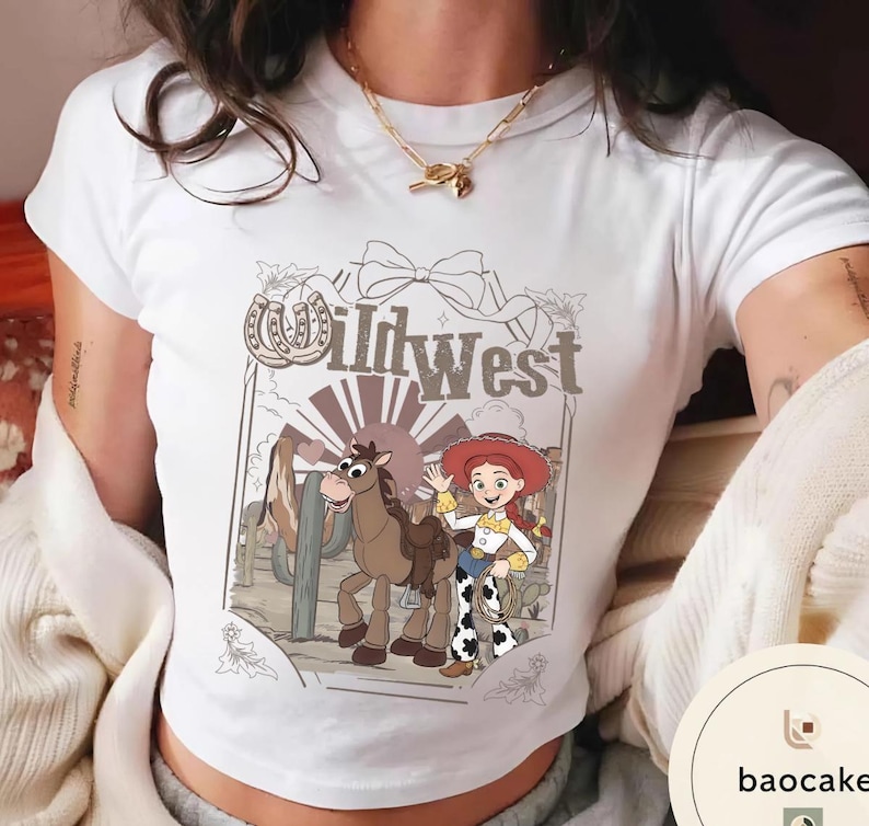 Pu&ograve; includere: T-shirt bianca con grafica a cartoni animati del West selvaggio. Il design include una cowgirl dei cartoni animati, un cavallo e la scritta "Wild West" in stile vintage. La maglietta ha un girocollo classico e maniche corte.