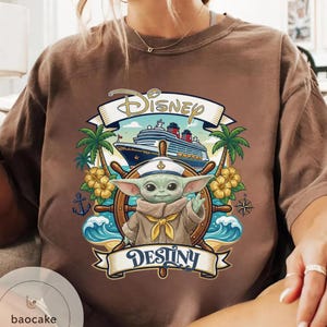 May include: Brown t-shirt featuring a graphic of Baby Yoda at the helm of a ship with the word "Disney" above and "Destiny" below. The design includes palm trees, waves, and a cruise ship.