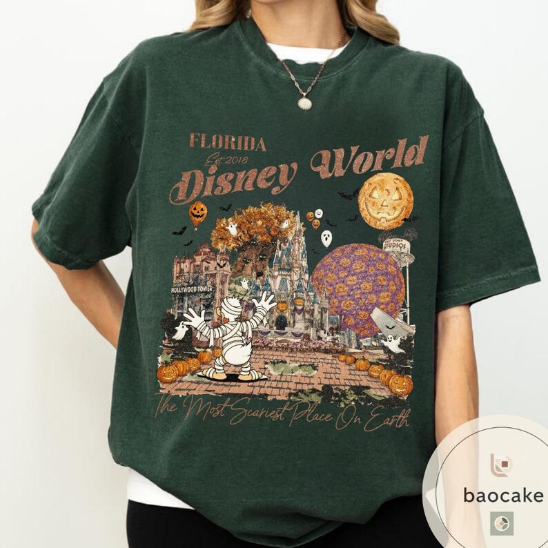 Op de afbeelding: Donkergroen T-shirt met een Halloween-thema Disney World graphic. Het ontwerp bevat een mummie, pompoenen, een spookkasteel en de tekst "Florida Disney World Est. 2018". Het shirt heeft ook de tekst "The Most Scariest Place On Earth".