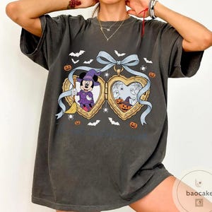 May include: Dark grey t-shirt with a heart-shaped locket design featuring Mickey Mouse in a witch costume and Halloween illustrations. The locket has a blue ribbon, bats, spiders, and pumpkins. The text "...a castle" is visible.