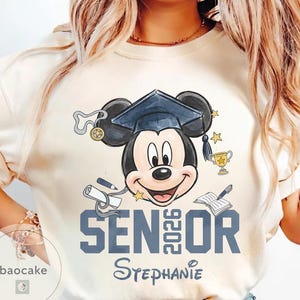 May include: Cream-colored t-shirt featuring Mickey Mouse wearing a graduation cap. The design includes the text "SENIOR 2026" in blue, along with a diploma, trophy, and stars. The name "Stephanie" is also printed on the shirt.