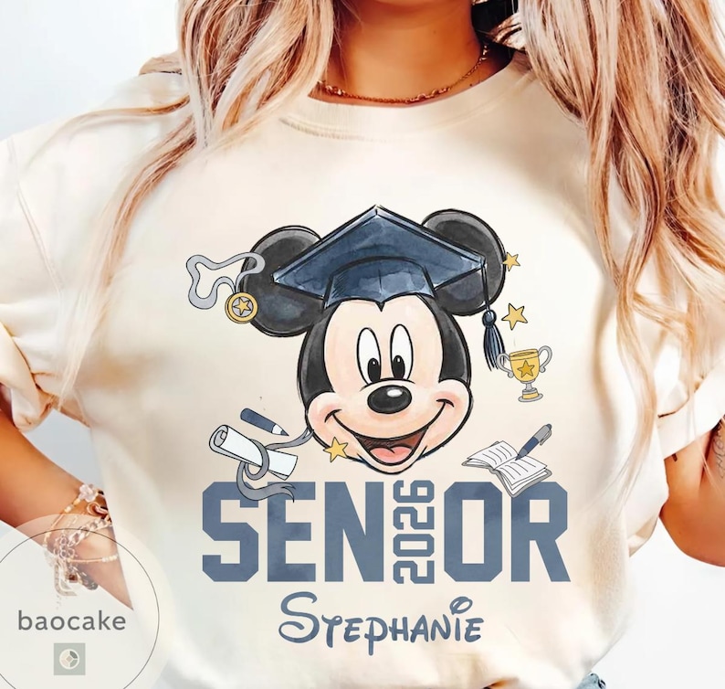 May include: Cream-colored t-shirt featuring Mickey Mouse wearing a graduation cap. The design includes the text "SENIOR 2026" in blue, along with a diploma, trophy, and stars. The name "Stephanie" is also printed on the shirt.