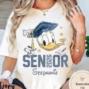 May include: Ivory t-shirt featuring a graphic of Donald Duck wearing a graduation cap. The text "SENIOR 2026 Stephanie" is printed in blue. Additional graphics include stars, a trophy, and a book.