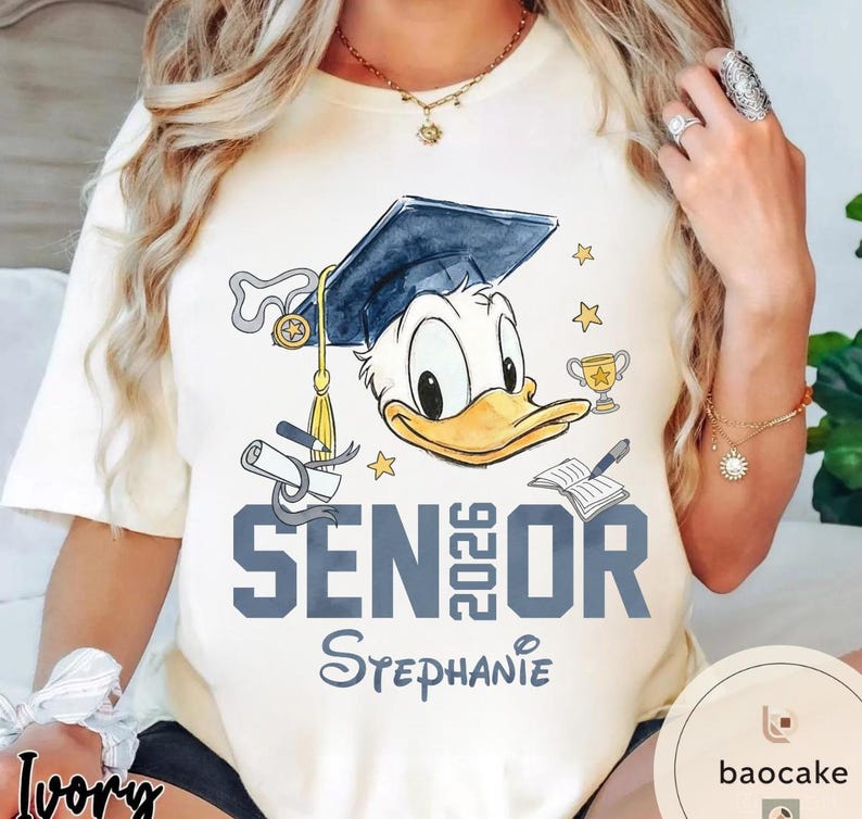 May include: Ivory t-shirt featuring a graphic of Donald Duck wearing a graduation cap. The text "SENIOR 2026 Stephanie" is printed in blue. Additional graphics include stars, a trophy, and a book.