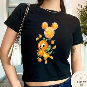 Op de afbeelding: Zwart T-shirt met een cartoon oranje vogel die een sinaasappel en een Mickey Mouse-vormige ballon vasthoudt. De vogel heeft groene bladeren en is omringd door sinaasappelschijfjes en kleine sinaasappels. Het shirt is een cropped model.