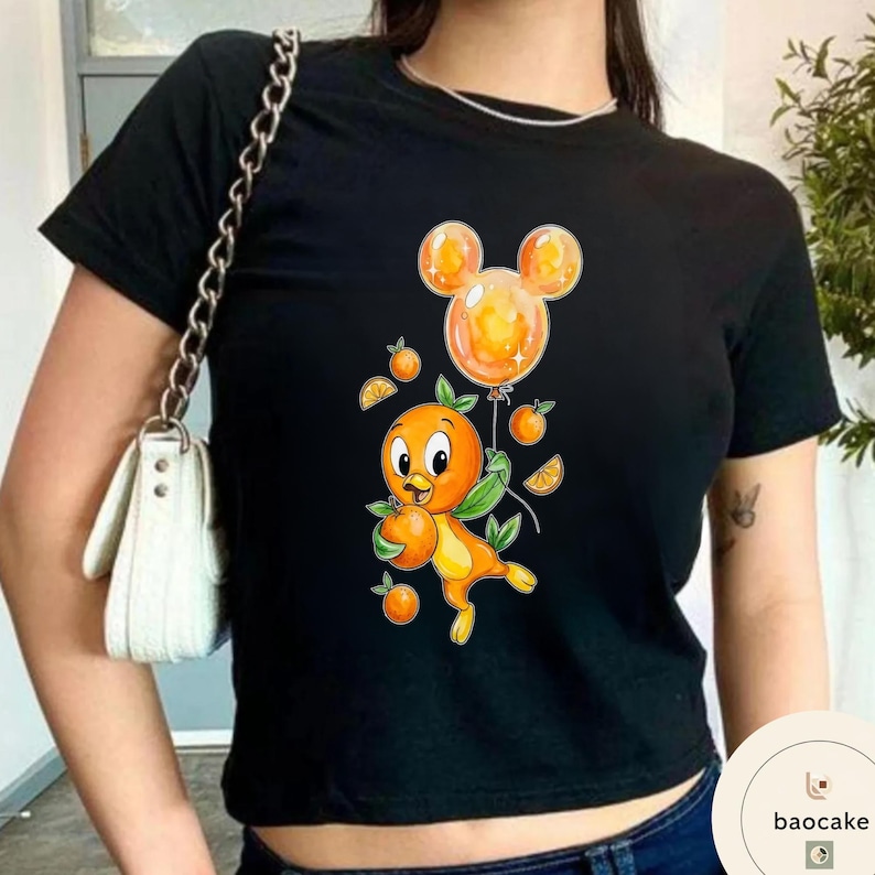 Op de afbeelding: Zwart T-shirt met een cartoon oranje vogel die een sinaasappel en een Mickey Mouse-vormige ballon vasthoudt. De vogel heeft groene bladeren en is omringd door sinaasappelschijfjes en kleine sinaasappels. Het shirt is een cropped model.
