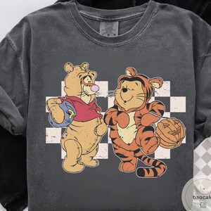 May include: Dark gray t-shirt featuring two cartoon tigers. One tiger wears a red hoodie and carries a blue bag. The other tiger is dressed in an orange tiger costume and holds a carved pumpkin. A white and gray checkered pattern is behind the tigers.