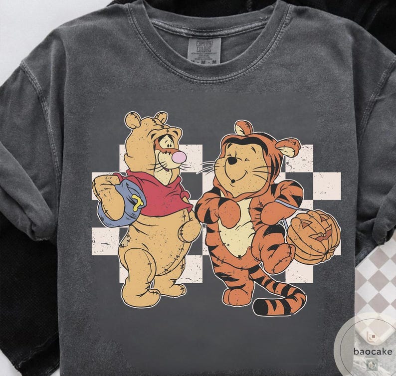 May include: Dark gray t-shirt featuring two cartoon tigers. One tiger wears a red hoodie and carries a blue bag. The other tiger is dressed in an orange tiger costume and holds a carved pumpkin. A white and gray checkered pattern is behind the tigers.