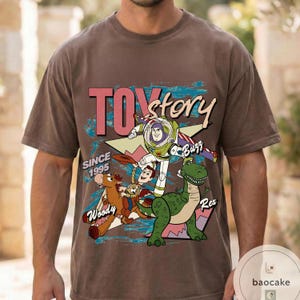 May include: Brown t-shirt featuring a graphic of characters from the movie "Toy Story." The design includes Buzz Lightyear, Woody, Rex, and the text "Toy Story" and "Since 1995." The shirt has a vintage, distressed look.