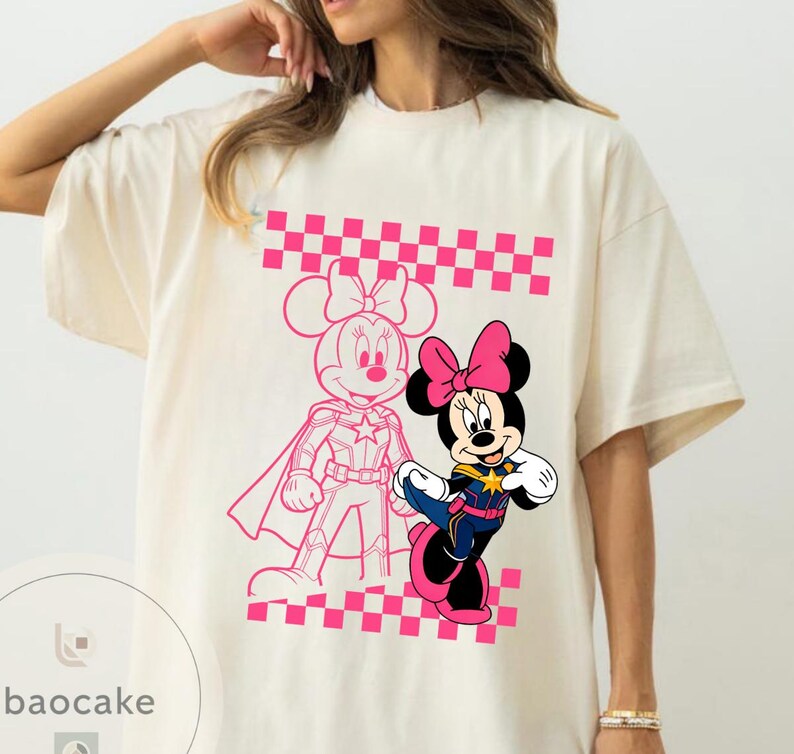 May include: Cream-colored t-shirt featuring a graphic of Minnie Mouse in a superhero outfit. The design includes a pink checkered pattern and a pink outline of Minnie Mouse in a superhero pose. The shirt has short sleeves.