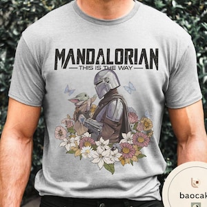 Puede incluir: Camiseta gris claro con un gr&aacute;fico de The Mandalorian y Grogu rodeados de flores y mariposas. El texto "MANDALORIAN - THIS IS THE WAY -" est&aacute; impreso en negro en la parte superior.