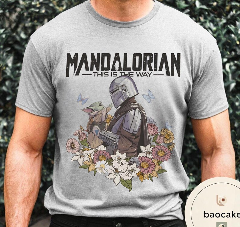 Puede incluir: Camiseta gris claro con un gr&aacute;fico de The Mandalorian y Grogu rodeados de flores y mariposas. El texto "MANDALORIAN - THIS IS THE WAY -" est&aacute; impreso en negro en la parte superior.
