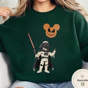 May include: Dark green sweatshirt featuring a Halloween-themed graphic. The design includes a Darth Vader character in a mummy costume holding a red lightsaber, with a Jack Skellington-inspired Mickey Mouse balloon. The sweatshirt has a crew neck.