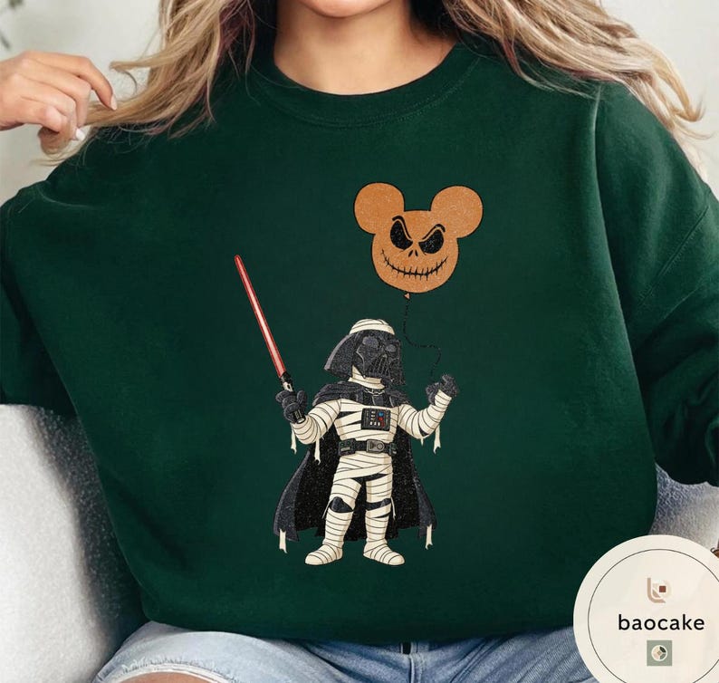 May include: Dark green sweatshirt featuring a Halloween-themed graphic. The design includes a Darth Vader character in a mummy costume holding a red lightsaber, with a Jack Skellington-inspired Mickey Mouse balloon. The sweatshirt has a crew neck.