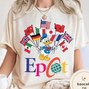 May include: A cream-coloured t-shirt with a cartoon Donald Duck holding flags from different countries. The word "Epcot" is printed in colourful letters below the character. The shirt has a round neck and short sleeves.