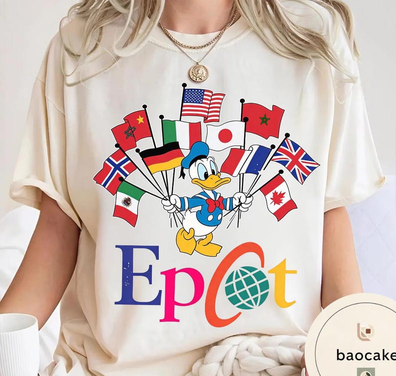 May include: A cream-coloured t-shirt with a cartoon Donald Duck holding flags from different countries. The word "Epcot" is printed in colourful letters below the character. The shirt has a round neck and short sleeves.
