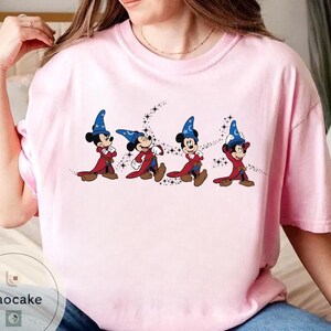 May include: A light pink t-shirt featuring a graphic of Mickey Mouse in a sorcerer's outfit. The cartoon character is depicted in a series of poses, wearing a blue hat with a crescent moon and stars, a red robe, and brown shoes.