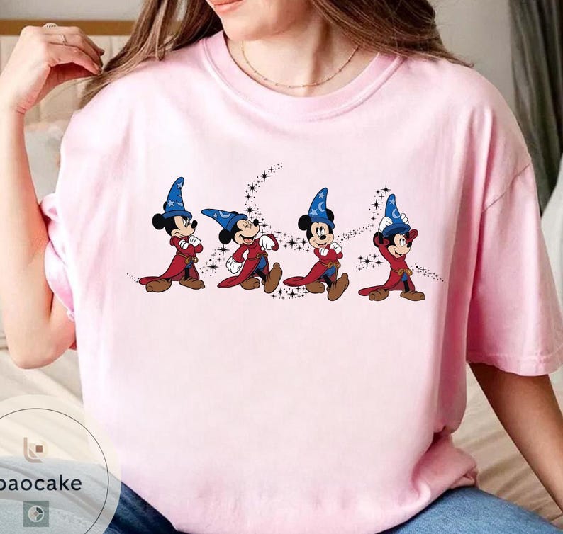May include: A light pink t-shirt featuring a graphic of Mickey Mouse in a sorcerer's outfit. The cartoon character is depicted in a series of poses, wearing a blue hat with a crescent moon and stars, a red robe, and brown shoes.