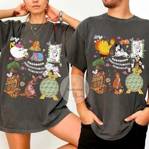 May include: Dark gray t-shirts with colorful cartoon character graphics. The designs include various characters and elements, such as a teapot, a kangaroo, and a building. The shirts are worn by two people.
