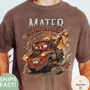 May include: Brown t-shirt with a cartoon image of Mater from the Cars film. The graphic includes the word "MATER" in a distressed font, with lightning bolts and tools. The tow truck Mater is smiling and has his headlights on.