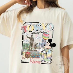 Puede incluir: Camiseta color crema con un gr&aacute;fico colorido de Tokyo Disneyland. El dise&ntilde;o incluye las palabras "TOKYO Disneyland" y una ilustraci&oacute;n de Mickey Mouse con un kimono sosteniendo un globo de arco&iacute;ris, con una estatua y un castillo en el fondo.