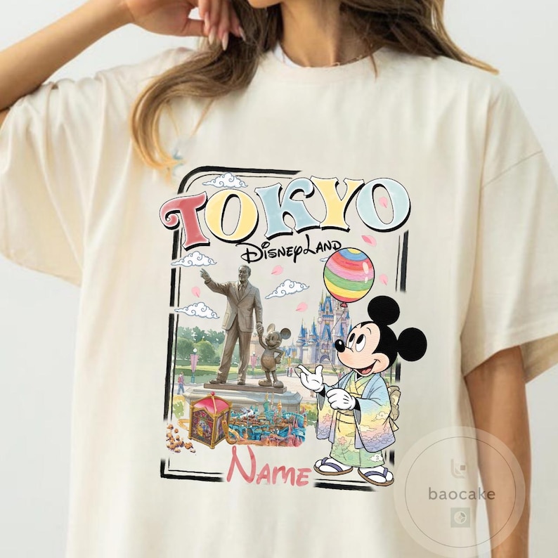 Puede incluir: Camiseta color crema con un gr&aacute;fico colorido de Tokyo Disneyland. El dise&ntilde;o incluye las palabras "TOKYO Disneyland" y una ilustraci&oacute;n de Mickey Mouse con un kimono sosteniendo un globo de arco&iacute;ris, con una estatua y un castillo en el fondo.