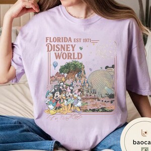 May include: A light purple t-shirt featuring a graphic print of Disney World landmarks and characters. The text reads FLORIDA EST 1971 DISNEY WORLD. The design includes Mickey Mouse, Minnie Mouse, Donald Duck, Daisy Duck, and Pluto, along with the Tree of Life and Epcot sphere.