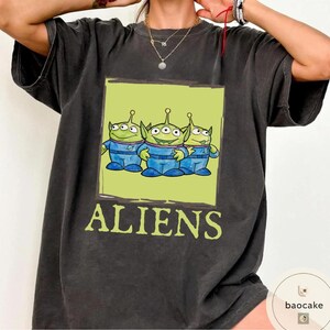 K&ouml;nnte beinhalten: Ein anthrazitfarbenes Kurzarm-T-Shirt mit einer rechteckigen Grafik von drei gr&uuml;nen, drei&auml;ugigen Au&szlig;erirdischen in blauen Raumanz&uuml;gen. Unter der Grafik ist das Wort ALIENS in hellgr&uuml;ner Schrift aufgedruckt. Das Shirt hat einen Rundhalsausschnitt.