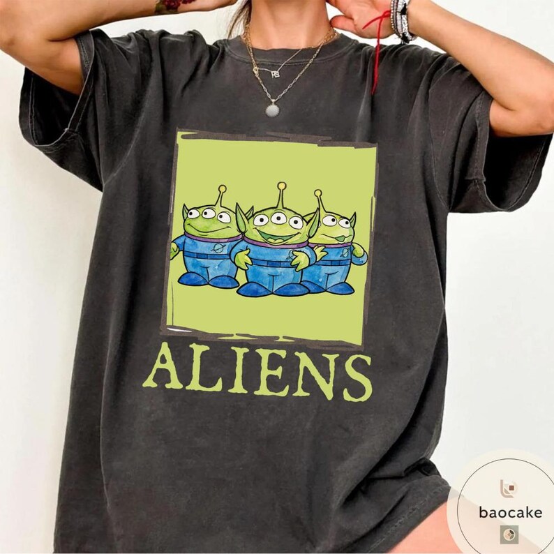 K&ouml;nnte beinhalten: Ein anthrazitfarbenes Kurzarm-T-Shirt mit einer rechteckigen Grafik von drei gr&uuml;nen, drei&auml;ugigen Au&szlig;erirdischen in blauen Raumanz&uuml;gen. Unter der Grafik ist das Wort ALIENS in hellgr&uuml;ner Schrift aufgedruckt. Das Shirt hat einen Rundhalsausschnitt.
