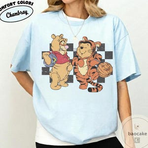 May include: Light blue t-shirt with a graphic of two cartoon characters in costumes. One is dressed as a tiger and the other as a bear. The shirt has the words "Comfort Colors" and "Chambray" in a circular design.