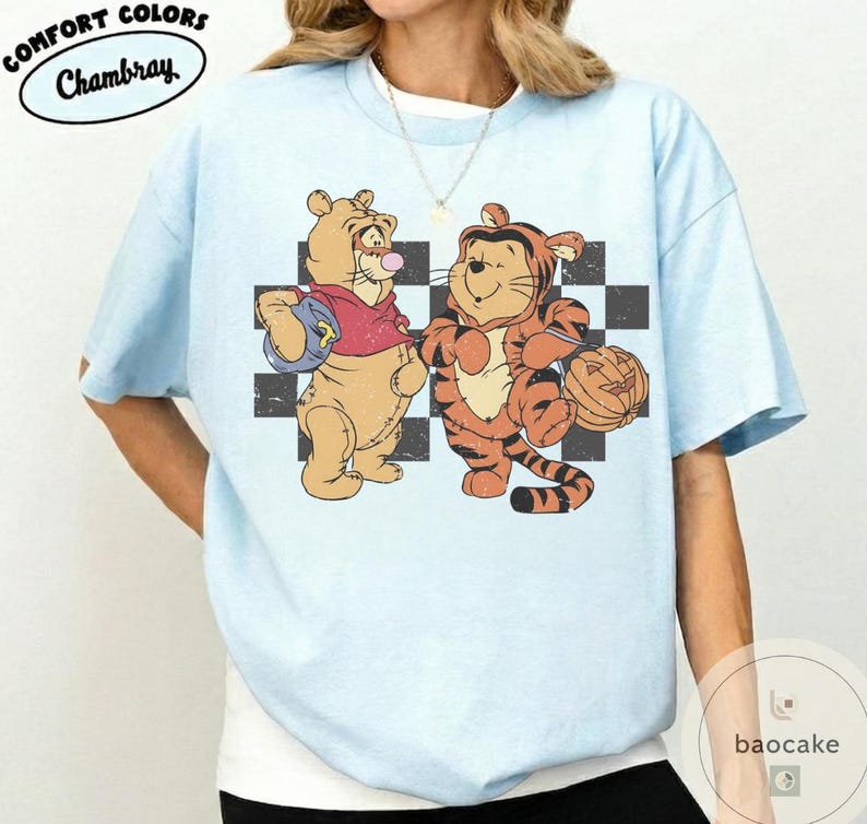 May include: Light blue t-shirt with a graphic of two cartoon characters in costumes. One is dressed as a tiger and the other as a bear. The shirt has the words "Comfort Colors" and "Chambray" in a circular design.