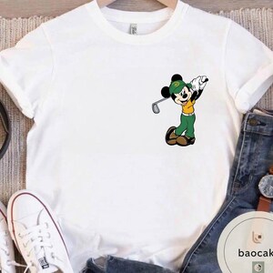 May include: White t-shirt featuring a cartoon Mickey Mouse character dressed in golf attire, swinging a golf club. Mickey is wearing a green hat, shirt, and pants with a yellow shirt. The shirt is on a hanger.
