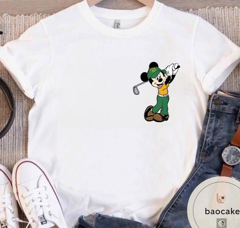 May include: White t-shirt featuring a cartoon Mickey Mouse character dressed in golf attire, swinging a golf club. Mickey is wearing a green hat, shirt, and pants with a yellow shirt. The shirt is on a hanger.