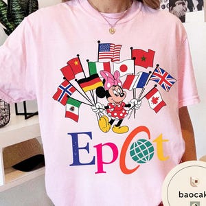 May include: A pale pink t-shirt with a Minnie Mouse graphic holding flags from different nations. Minnie is in a red polka dot dress and pink bow. The word "Epcot" is printed in blue, orange, and yellow.
