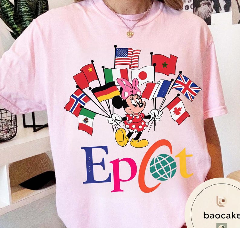 May include: A pale pink t-shirt with a Minnie Mouse graphic holding flags from different nations. Minnie is in a red polka dot dress and pink bow. The word "Epcot" is printed in blue, orange, and yellow.