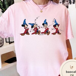 May include: Light pink t-shirt featuring a graphic of Mickey Mouse in a sorcerer's outfit, with a blue hat and red robe. The design includes a starry trail behind each character. The shirt is a casual wear item.