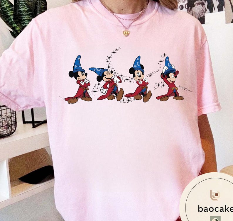 May include: Light pink t-shirt featuring a graphic of Mickey Mouse in a sorcerer's outfit, with a blue hat and red robe. The design includes a starry trail behind each character. The shirt is a casual wear item.