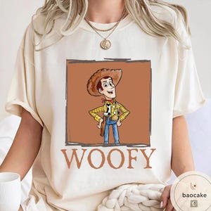 K&ouml;nnte beinhalten: Ein cremefarbenes Kurzarm-T-Shirt mit einer Grafik eines Cartoon-Cowboy-Charakters, der einen braunen Hut, ein gelbes kariertes Hemd und eine blaue Hose tr&auml;gt, in einem braunen quadratischen Rahmen, mit dem Wort WOOFY in gro&szlig;en braunen Buchstaben.